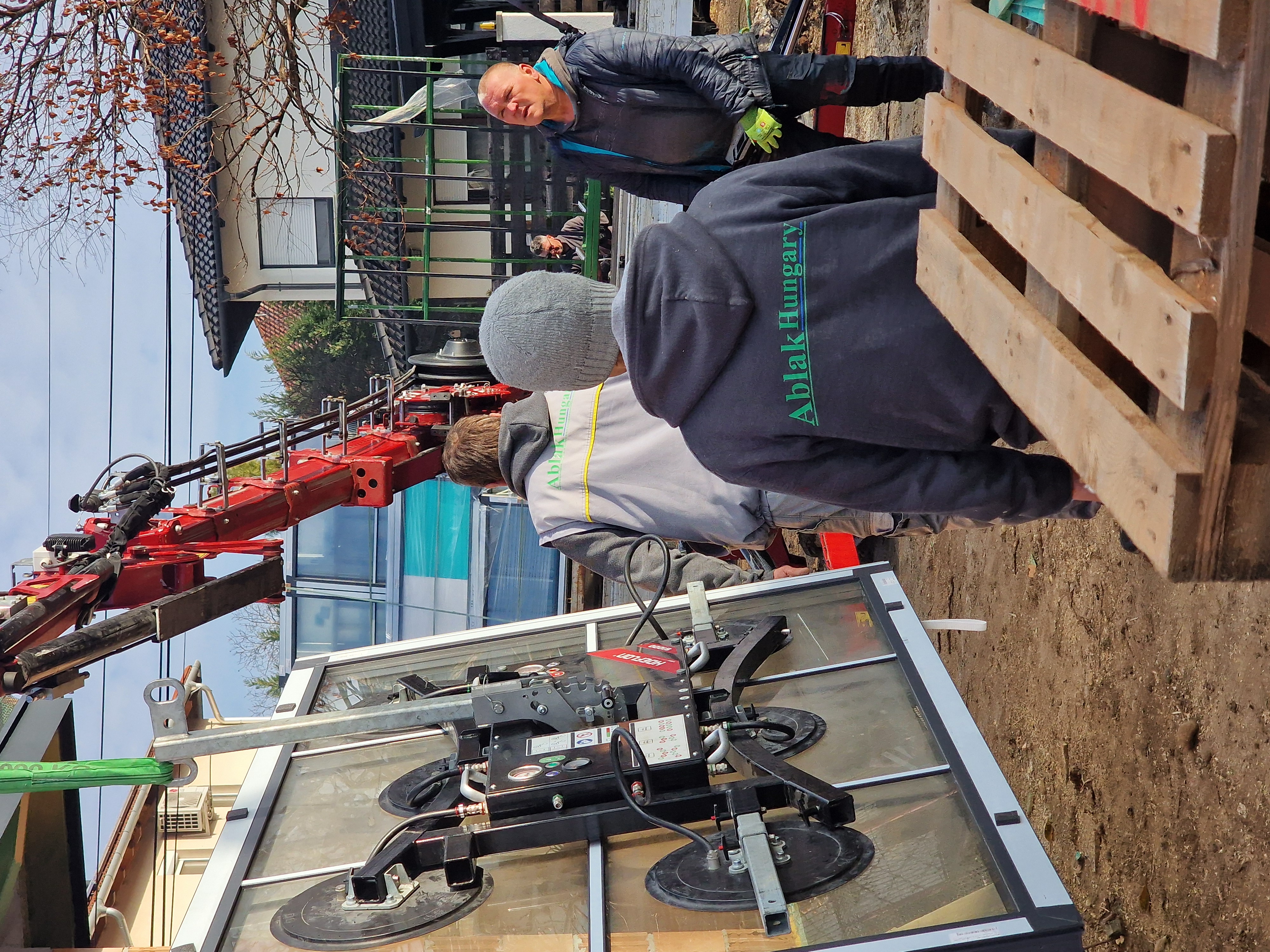 Munkások ablakot szerelnek be egy piros daru segítségével, AblakHungary feliratú ruhában.